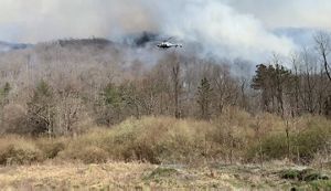 A white helicopter flies over a forest during a controlled burn. White smoke rises from the forest floor. The terrain rises into a ridge top in the background.