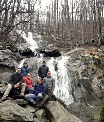 Group photo of the six members of the Allegheny Highlands Program team sitting on a rock outcrop. A waterfall cascades in two steps over the rocks behind them.