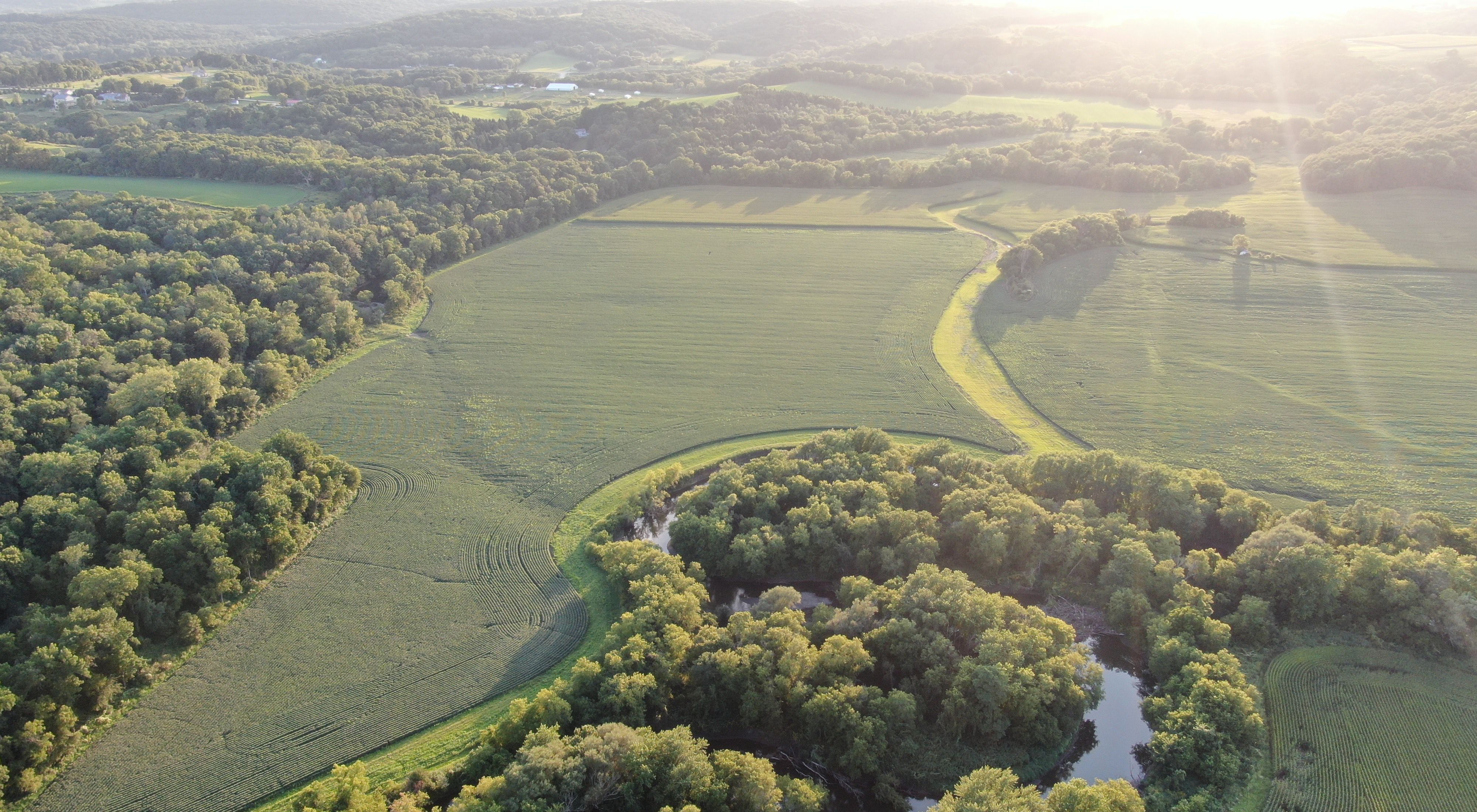 Aerial view of a river, crop fields and forest with the sunset in the background.
