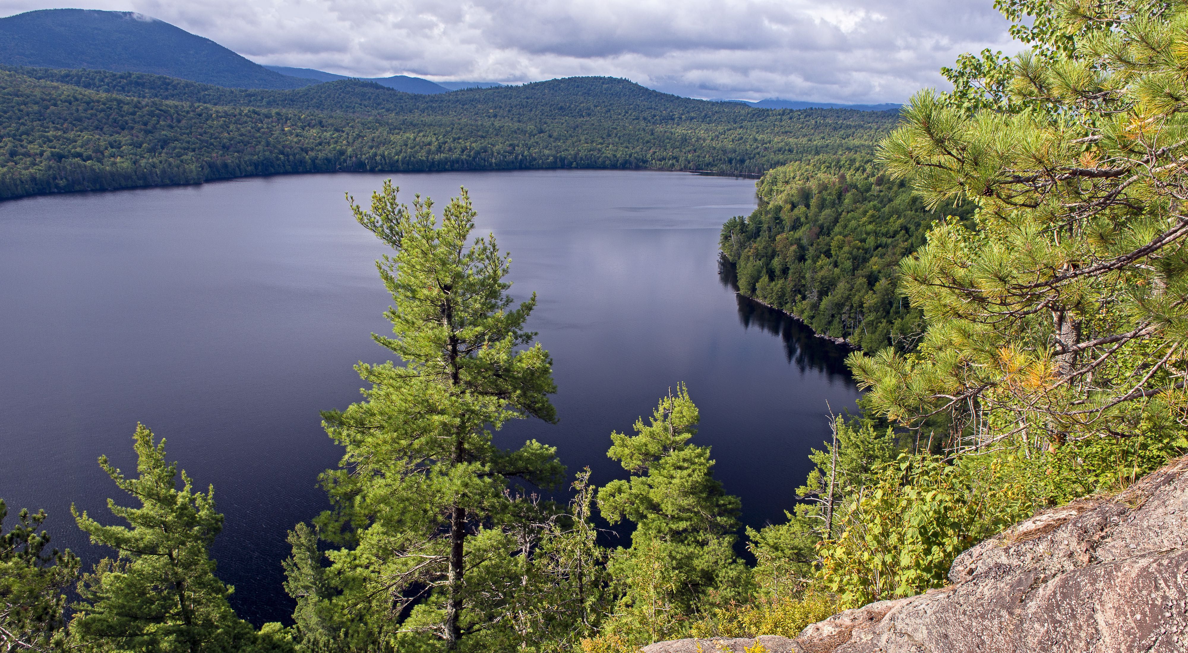 A view from atop a gray boulder on the righthand side of the image looking out onto a blue lake with green trees lining the shore.