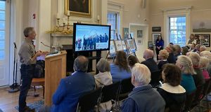 Joshua Royte stands at a podium and next to a video screen presenting to an audience.