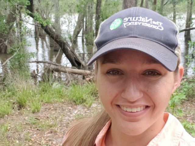 A selfie of Julia wearing a TNC baseball cap in front of a wetland