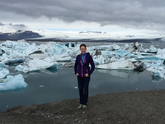 Kathleen on a black sandy shore with ice bergs in the water behind her.