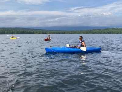 Three kids paddle kayaks of different colors in a lake.
