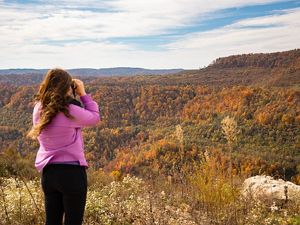 A woman stands on a ridge overlooking a valley and hills on the other side while looking through binoculars to behold Kentucky's natural beauty.