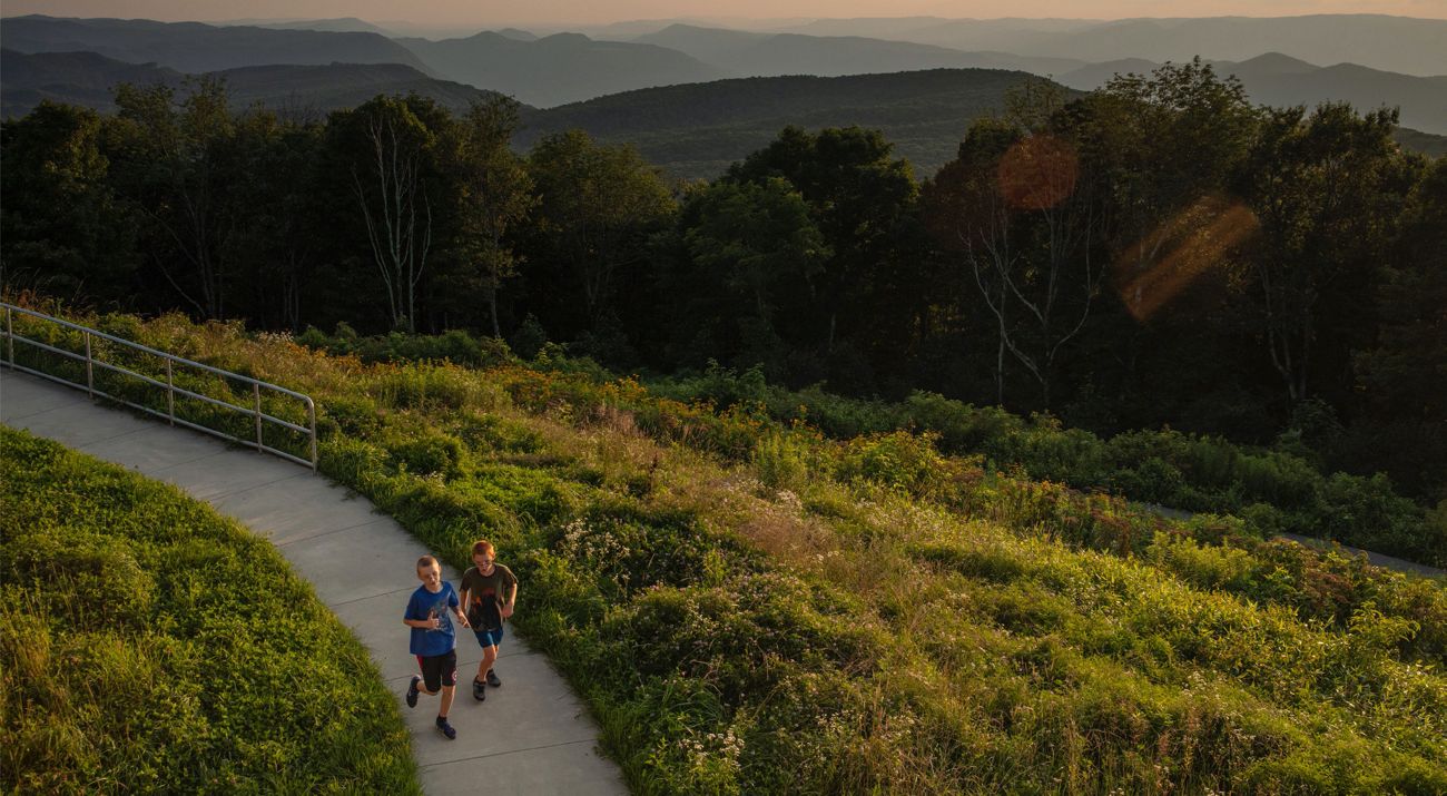 Two children run down a concrete path surrounded by grasslands and forest.
