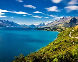 High-altitude view looking across the bright blue waters of Lake Wakatipu and the road from Queenstown to Glenorchy in New Zealand; southern Alps mountains are in the distance.