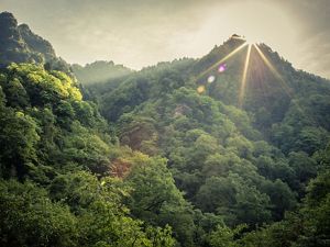 The sun peeks out from behind mountains covered in lush forests.