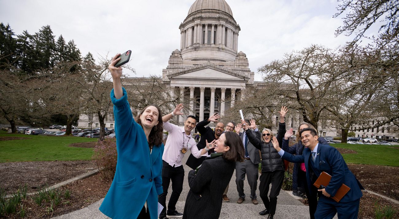 A group of people take a selfie and wave at the smartphone camera as they stand in front of the WA state capitol building.