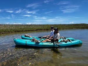 Two women in blue kayaks sit in shallow water above an oyster reef restoration site in North Carolina.