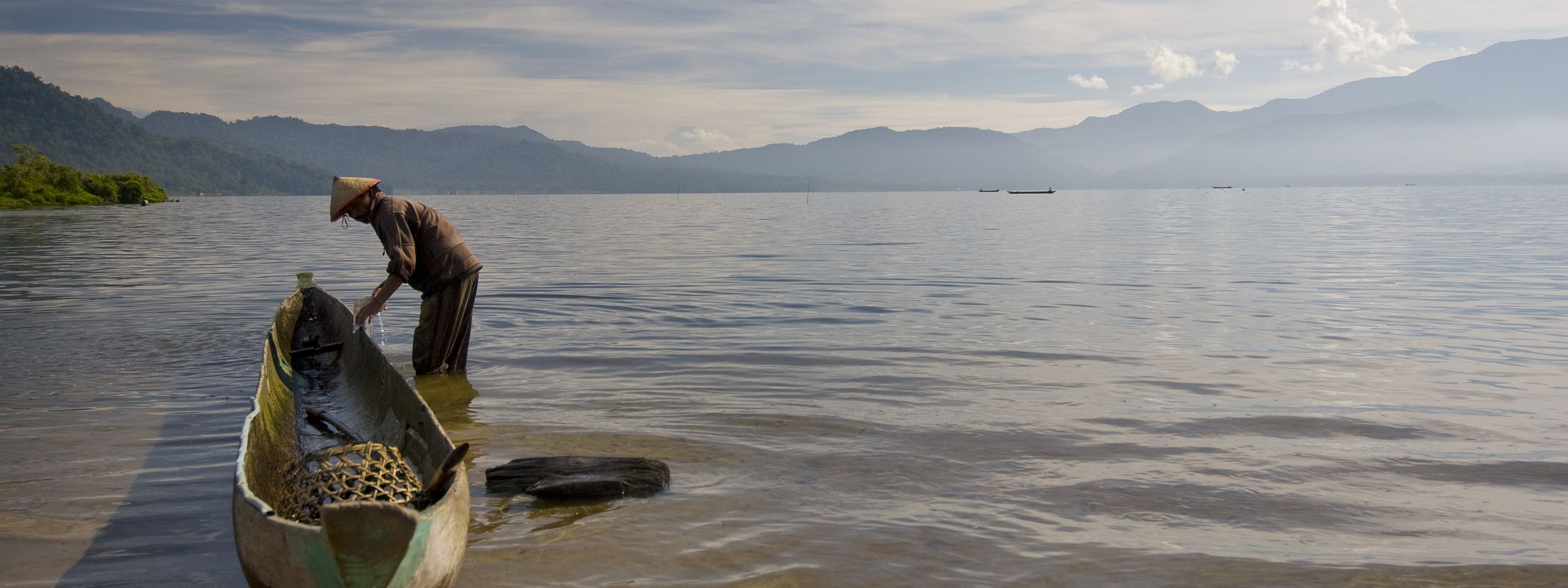 A man stands in knee-deep water next to a dugout canoe; mountains surround the lake in the distance.