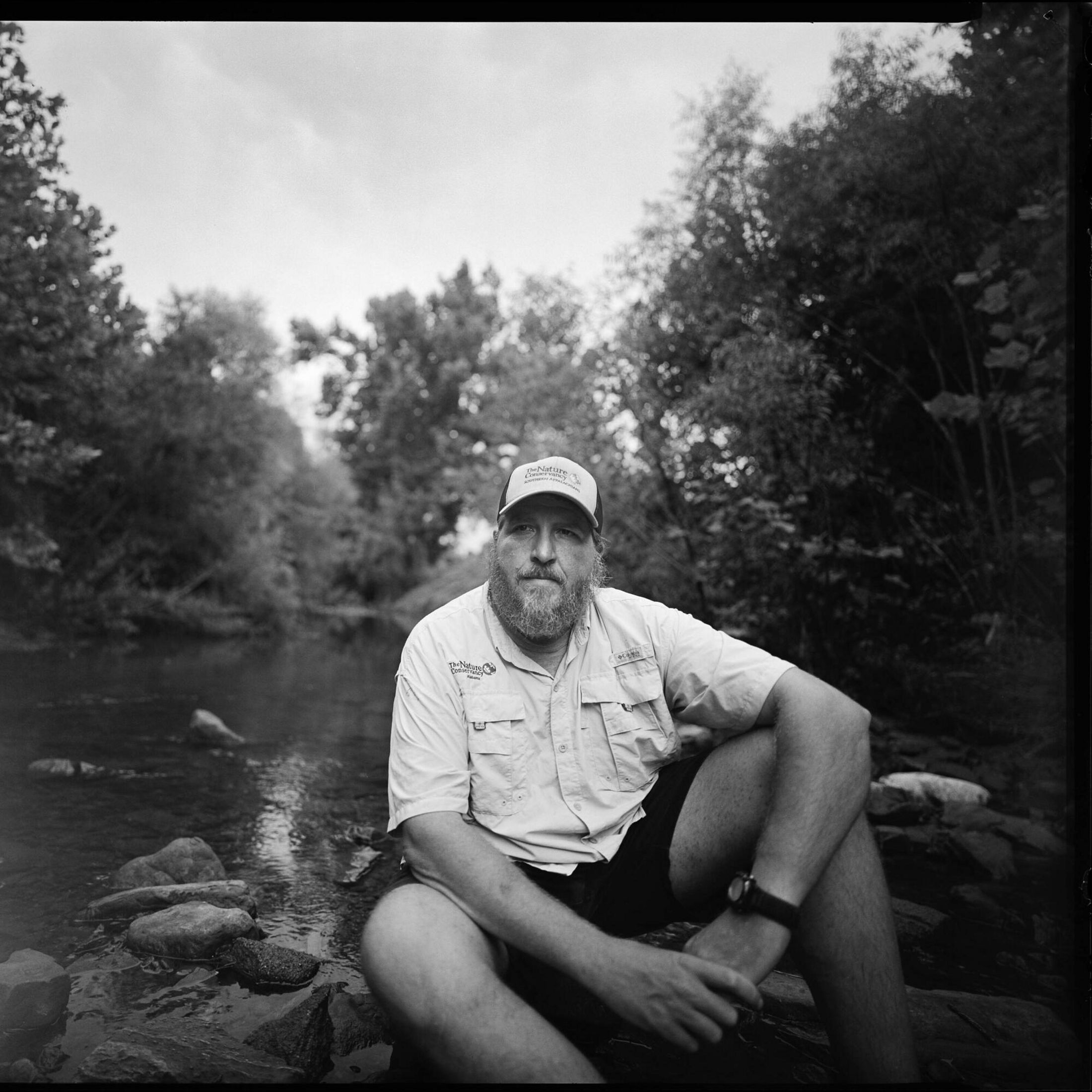 Black-and-white image of Jason Throneberry sitting at the edge of a river.