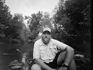 Black-and-white image of Jason Throneberry sitting at the edge of a river.