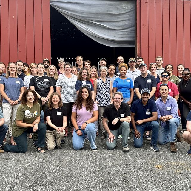 Group photo of TNC MD/DC staff. A large group of people stand together outdoors in front of a red barn, smiling during the chapter's annual staff retreat.