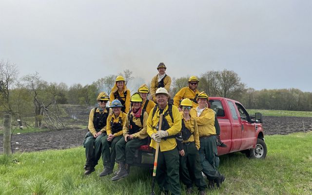 A group of land managers gather at the bed of a truck that is resting in front of an area where a prescribed burn was conducted. They are all wearing protective gear. 