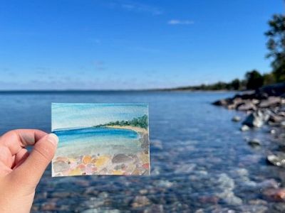 A hand holds a watercolor of Lake Superior in front of Lake Superior. 