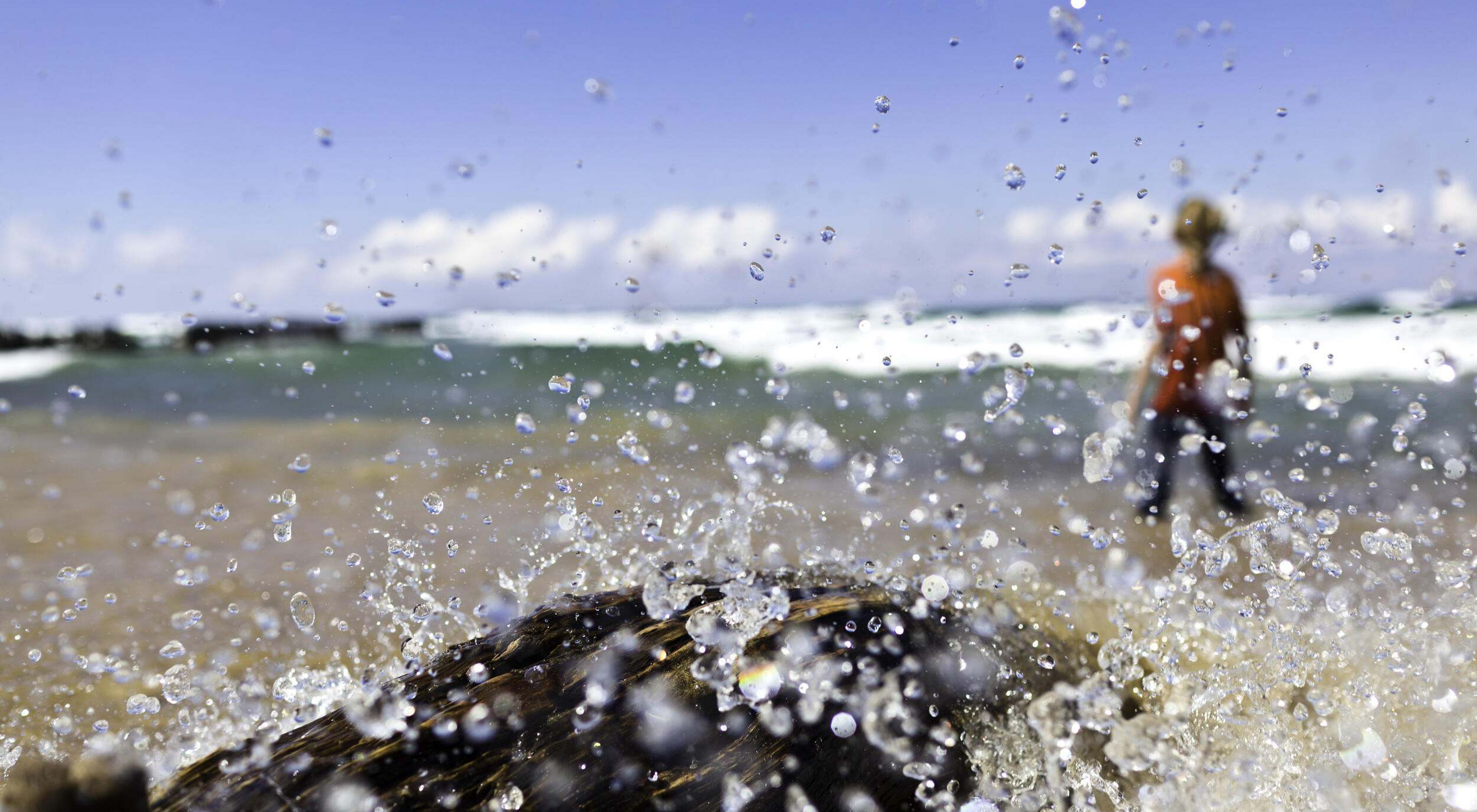 Water splashes over a rock in the foreground of the sandy shore of a lake with a person out of focus in the background. 