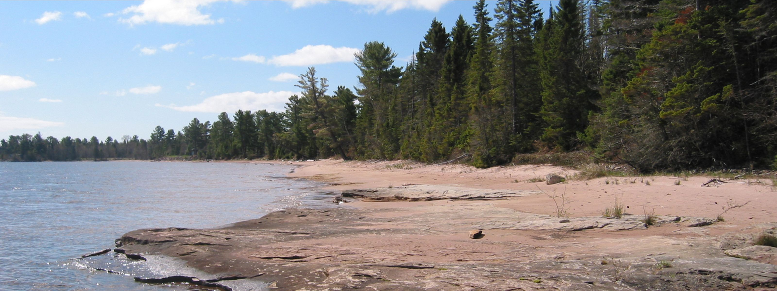 View of a rocky and sandy shoreline of a large body of water with forests along its banks.