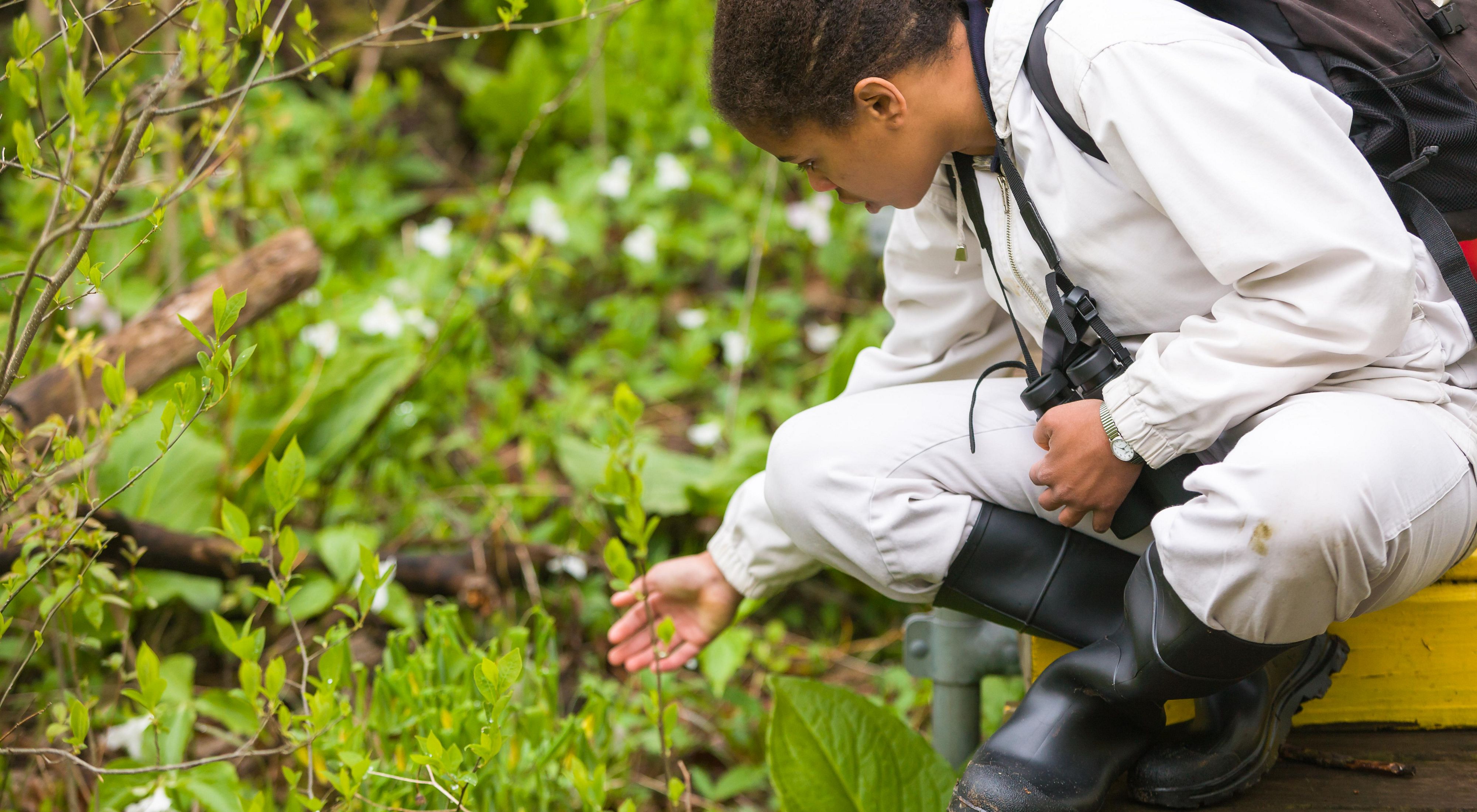 A girl wearing rubber boots and carrying binoculars bends over to look closely at a plant.