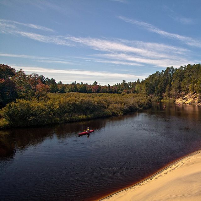 Kayakers paddle through the Two Hearted River. On the right is a sandy shore. On the left is a colorful forest of trees. Text overlay that says: 2021 Michigan Conservation Results Report.. 