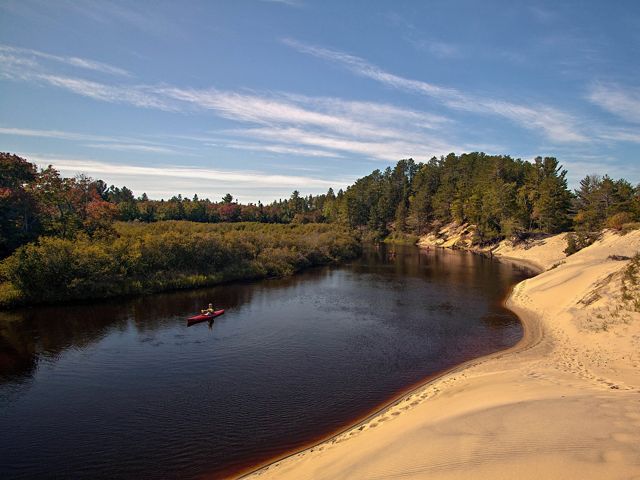 A few kayakers paddle along a winding river with a wide beach area on one bank and thick woods on the other bank.