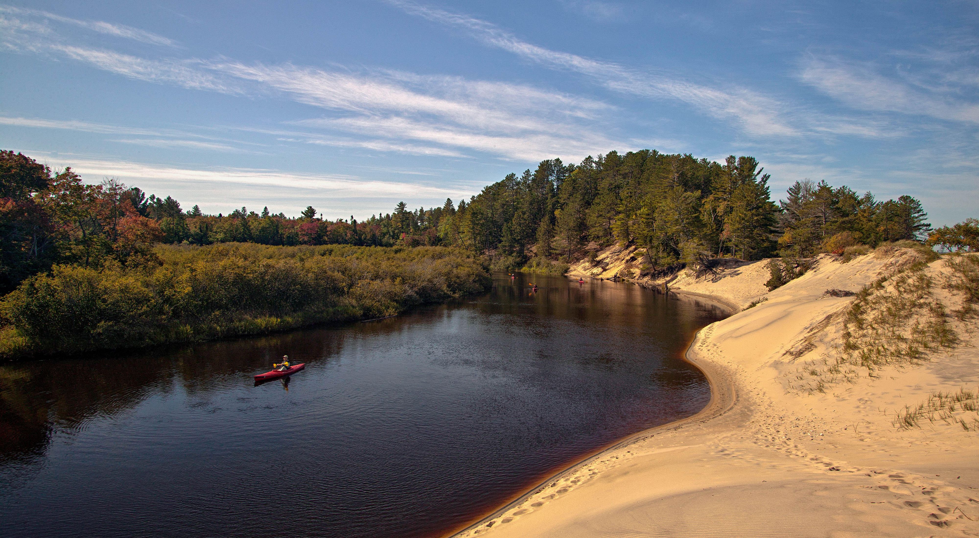 Kayakers paddle through the Two Hearted River. On the right is a sandy shore. On the left is a colorful forest of trees. 
