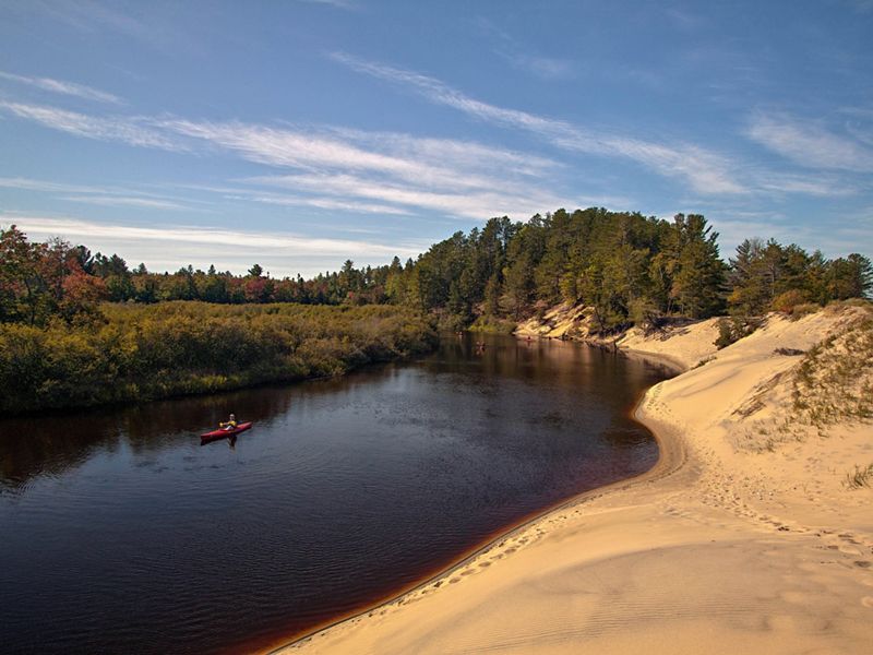 People kayak down a bend of the Two-Hearted River in Michigan. On the left of the river is autumn foliage and on the right is a sandy shore.