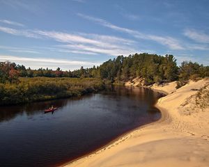 People kayak down a bend of the Two-Hearted River in Michigan. On the left of the river is autumn foliage, and on the right is a sandy shore.