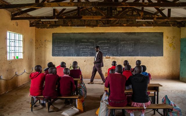 a man stands at the front of a classroom and a group of students in red clothing look up at him and a chalkboard
