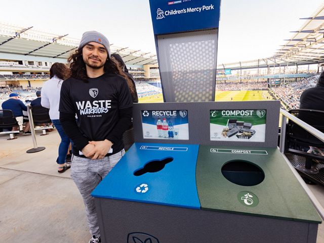 A man standing next to a recycle and compost bin at the soccer stadium.