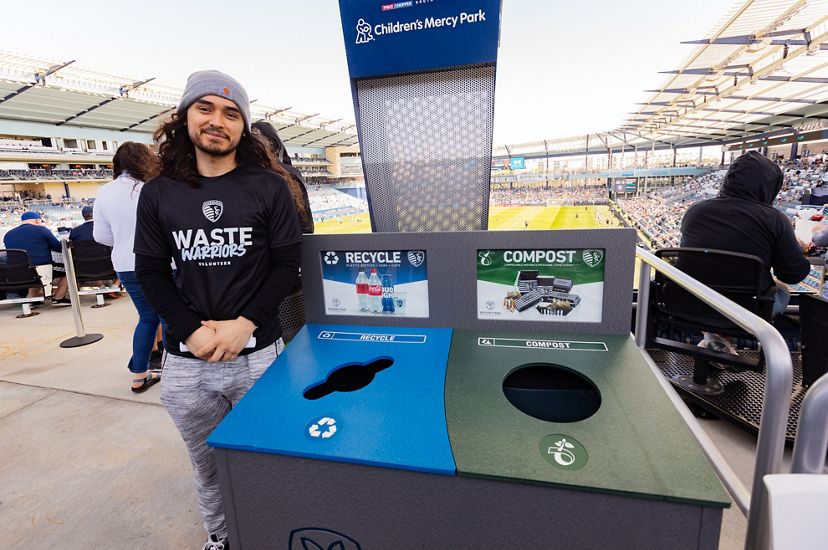 A man standing next to a recycle and compost bin at the soccer stadium.