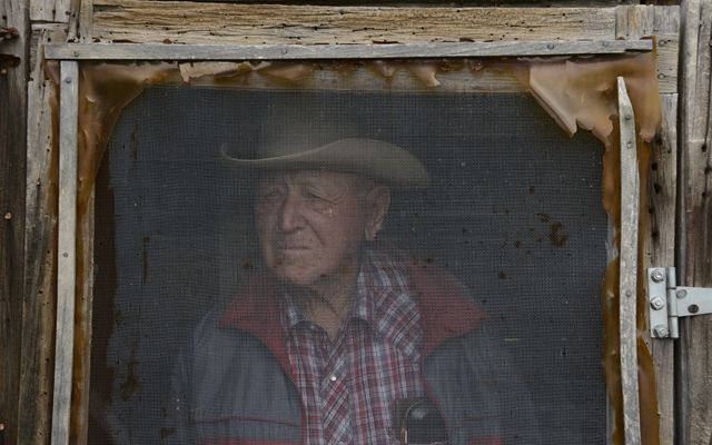 A weathered rancher in plaid stares out his screened window.