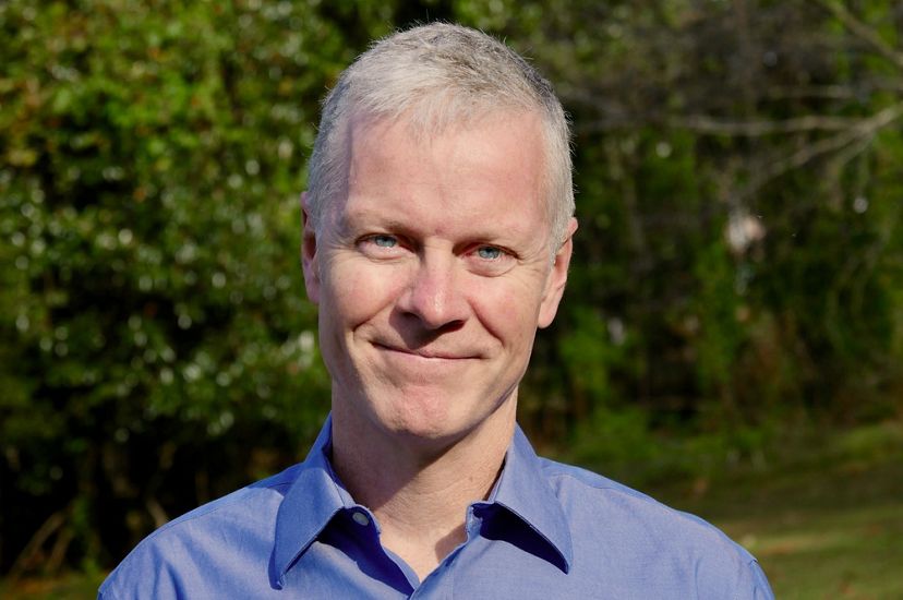 Close cropped head shot of a blond man wearing a blue shirt standing in front of green trees.