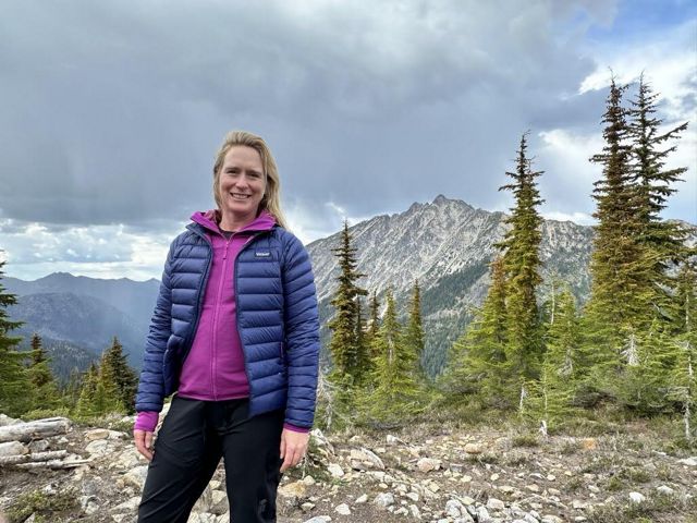 A woman stands in a rocky clearing. A sharp mountain peak rises behind her.