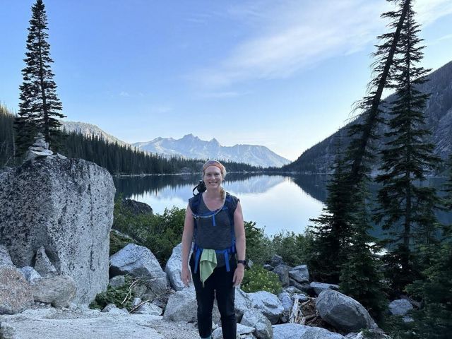 A woman stands in front of a mountain lake. The still surface of the water reflects the towering mountain peaks around it.