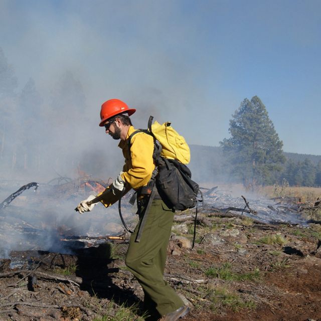 A person dressed in fire-protective clothing uses a hose to suppress a controlled burn near a pile of smoldering branches.