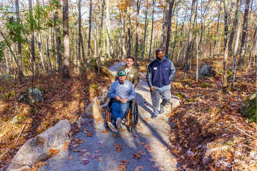 Un hombre y su amigo empujan a otro hombre en silla de ruedas por un camino llano y sinuoso en el bosque.