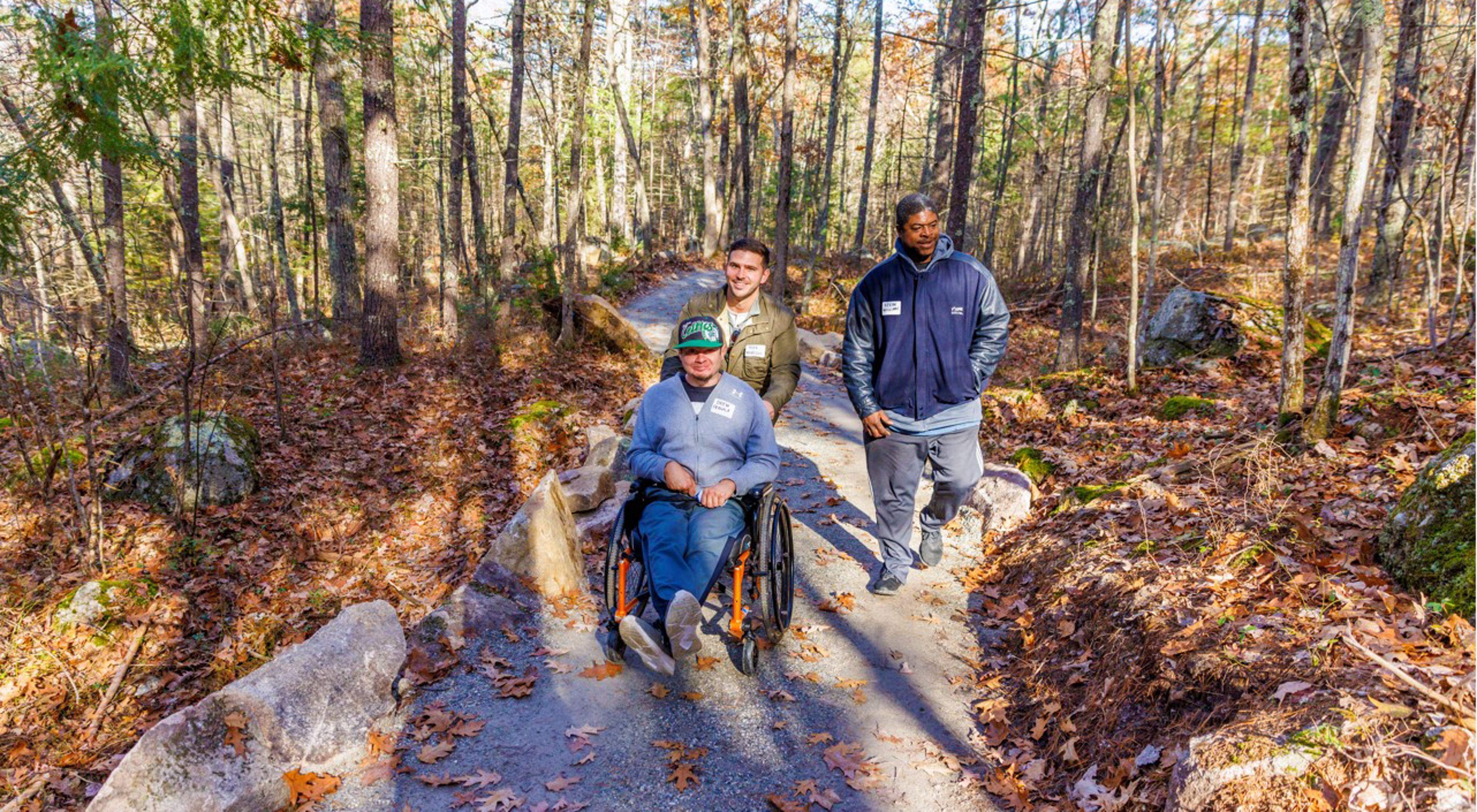 A man and his friend push another man using a wheelchair down a flat, winding path in the woods.