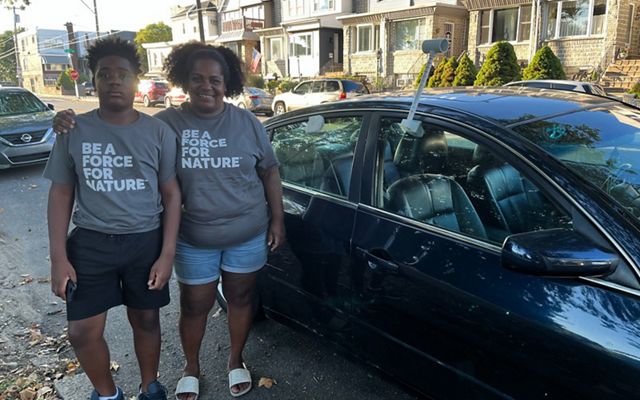 A woman and her son pose in gray shirts that read "be a force for nature" in front of a car. 
