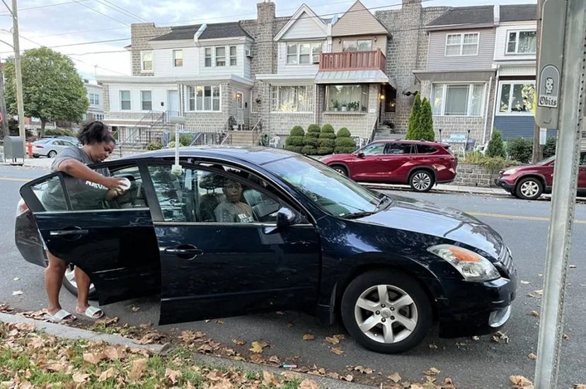 A car is parked on the street of a city neighborhood. A woman stands outside the car affixing a heat sensor to the rear passenger window.