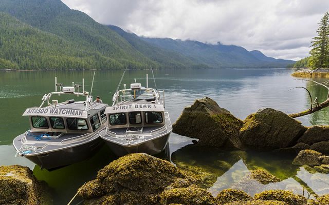 a boat named 'kitasoo watchman' is moored next to a boat named 'spirit bear 1' on a rocky coast with blue water and emerald mountains in the distance