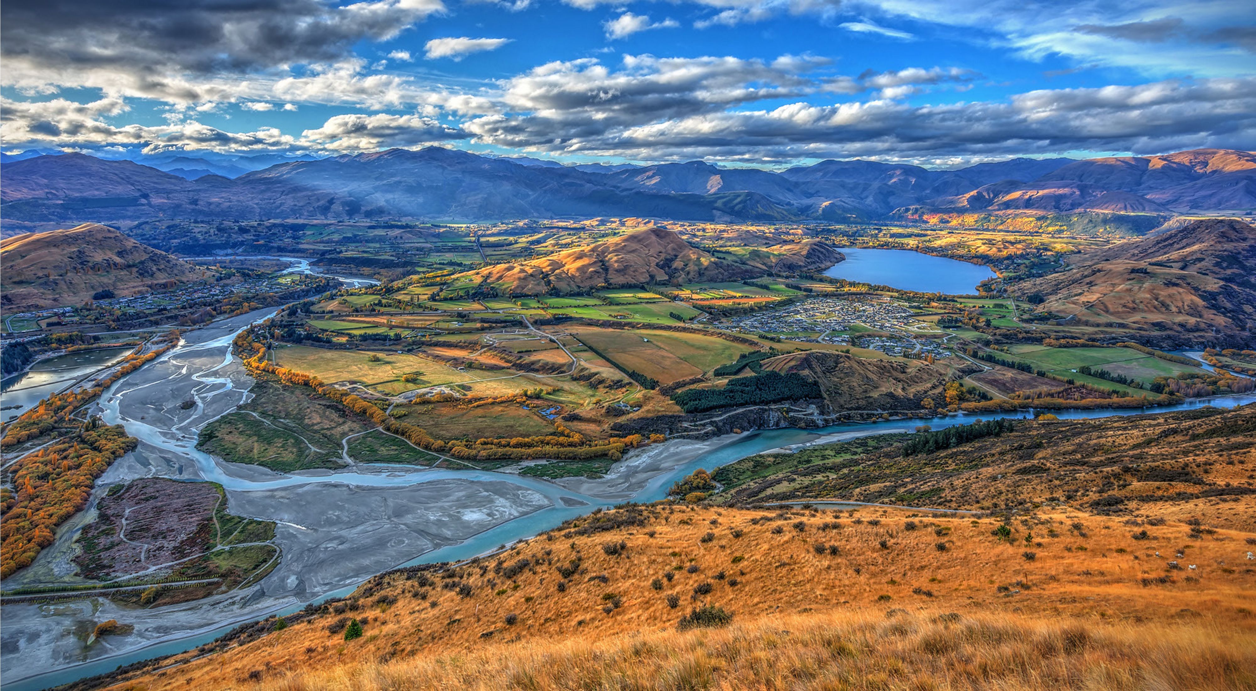 Aerial image of a wide expanse of golden mountains and snaking rivers under a blue sky filled with puffy white clouds in New Zealand.