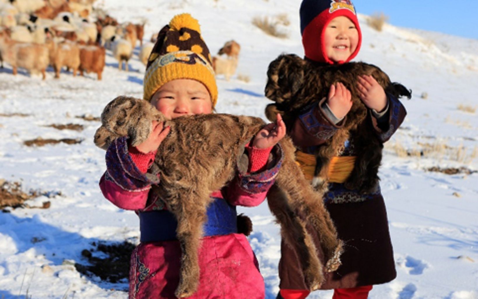 Two young Mongolian children stand in the snow while each holding a baby goat.