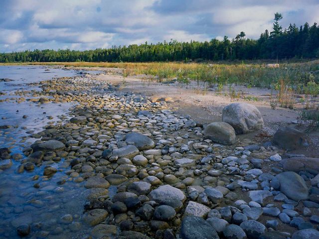 The bright, blue water of Lake Huron from the shore of the North Point Peninsula in Michigan. The shore is covered in small to large rocks and green foliage.
