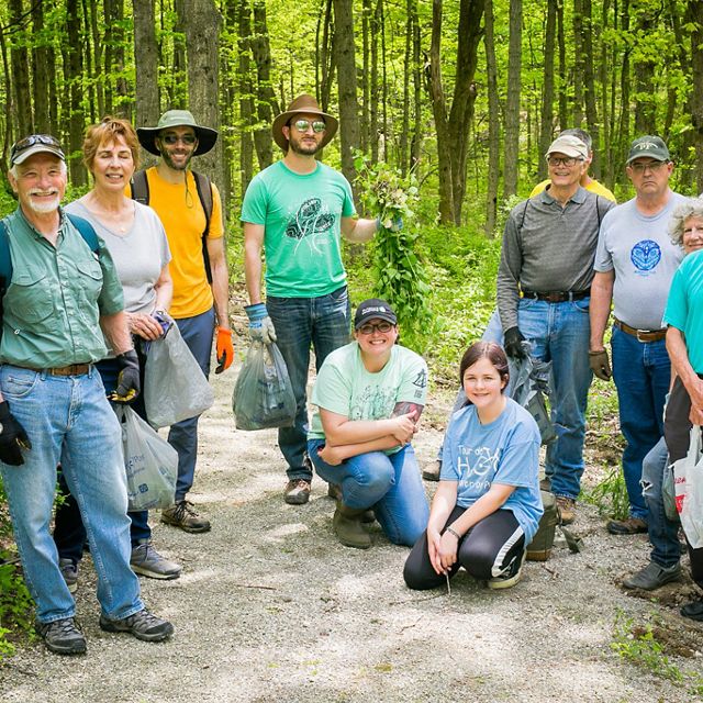 Group of smiling people stand in forest with bags of recently pulled invasive plants.
