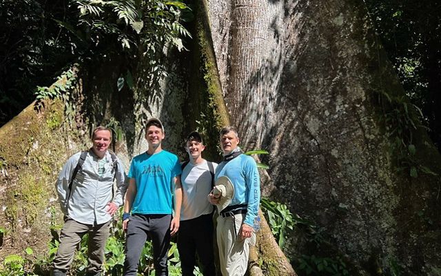 Chad Duplain with partner Matthew stand with two men in front of large tree in the Peruvian Amazon. 