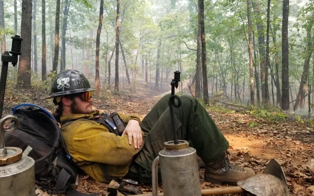 Man sits under a tree next to a drip torch during a controlled burn.