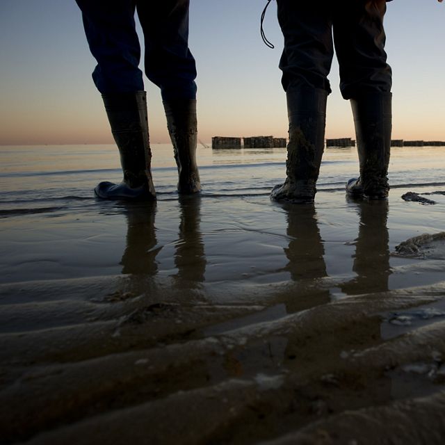 Beach-level view of the boots and lower legs of three people standing at the edge of a body of water.