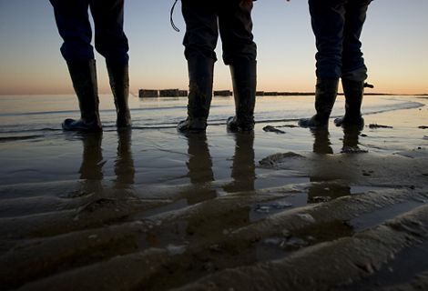 Beach-level view of the boots and lower legs of three people standing at the edge of a body of water.