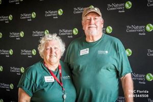 A man and woman wear green volunteer shirts and pose for a photo in front of a black TNC step-and-repeat banner.